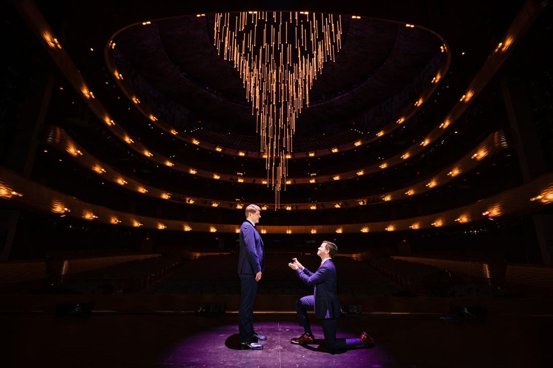 Man kneeling to propose to his partner on an empty opera house stage beneath a golden chandelier