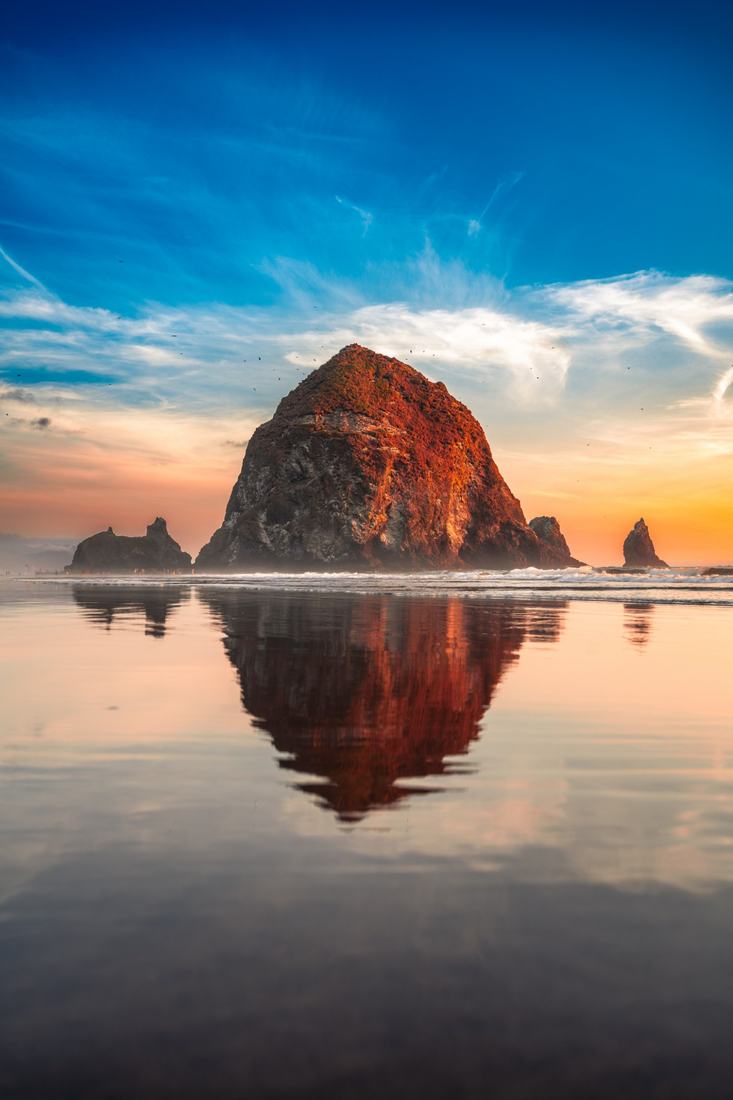 Haystack Rock glowing red at sunset, mirrored in wet sand at Cannon Beach