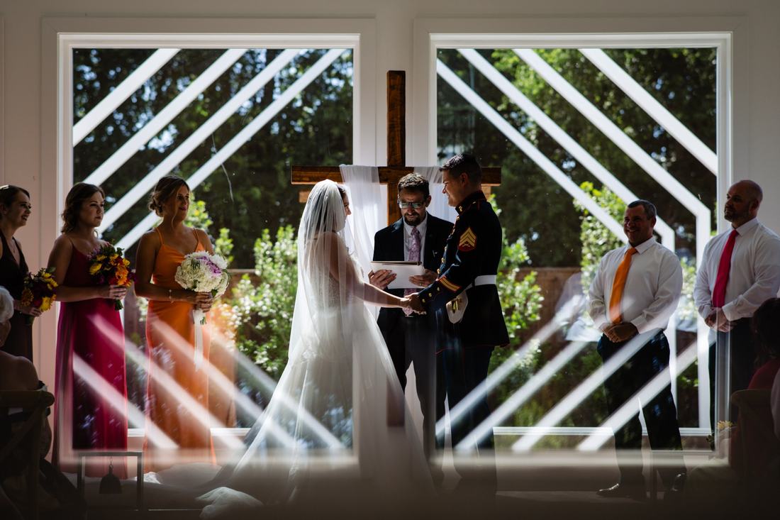 Bride and groom exchanging vows framed by geometric windows, with the wedding party on either side
