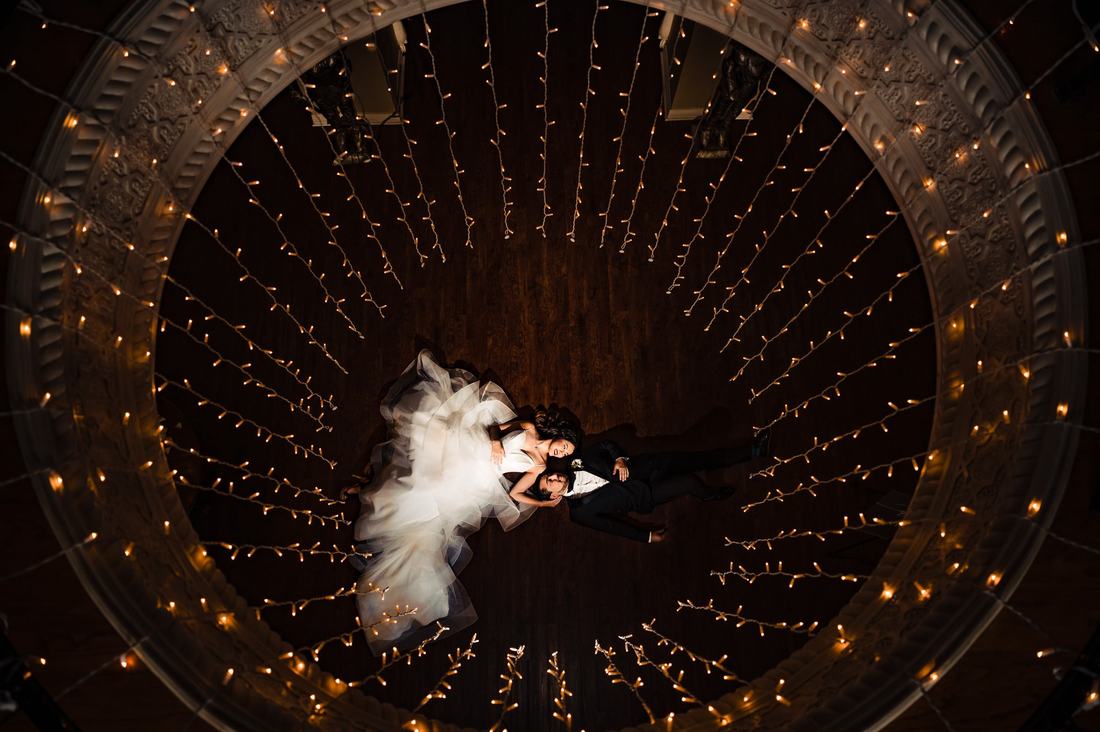 Overhead photo of a bride and groom lying on a floor surrounded by radiating string lights